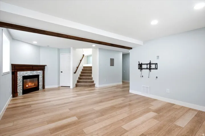 Wood-finish flooring, stone-surround fireplace with a dark wood mantle, exposed wood beam, recessed lighting, and carpeted staircase