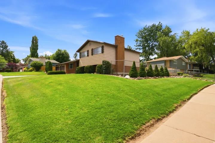 Front of home exterior featuring a brick chimney, light-toned siding, mature landscaping with shrubs, and a concrete sidewalk