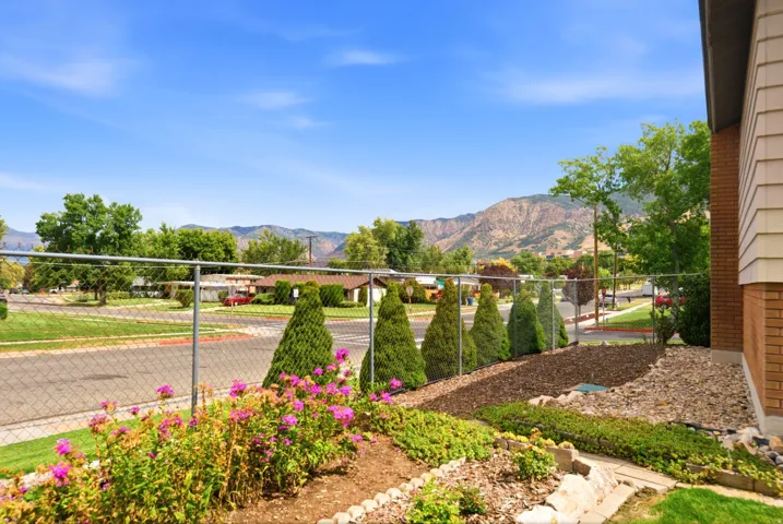 West exterior end of fully fenced backyard overlooking manicured landscaping, featuring evergreens, beautiful flowers, and views of the mountains