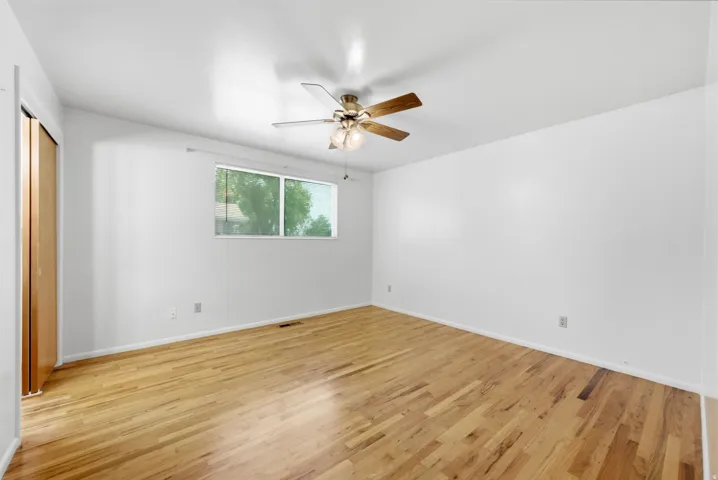 Upper level primary bedroom wth hardwood flooring, white walls, wood closet doors, ceiling fan with integrated lighting, window and blinds
