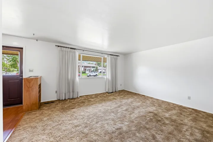Spacious front living room featuring neutral-toned carpet flooring, large window with sheer curtains allowing natural light, and white walls