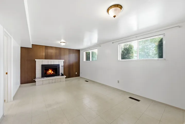 Lower level family room with wood paneling accent wall, white brick fireplace with hearth, two windows, vinyl tile flooring, and overhead lighting
