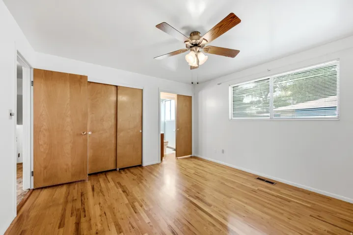 Upper level primary bedroom with hardwood flooring, ceiling fan with integrated lighting, wood closet doors, large window, three-quarter bathroom access, and blinds