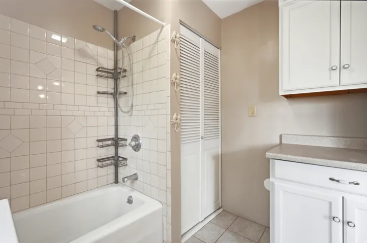 White tile shower surround with decorative inset, white bathtub, bi-fold louvered door, upper white cabinetry, and lower white cabinetry with a countertop