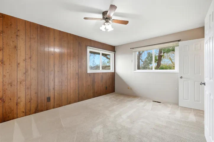 Wood-paneled accent wall, neutral wall paint, two windows, ceiling fan with light fixture, and carpeted flooring