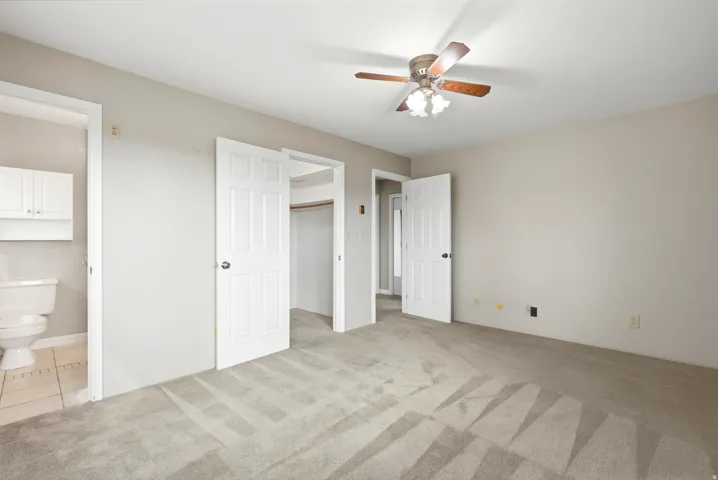 Neutral tone wall paint, light-colored carpeting, white six-panel doors, ceiling fan with light fixture, and an attached bathroom with tiled flooring