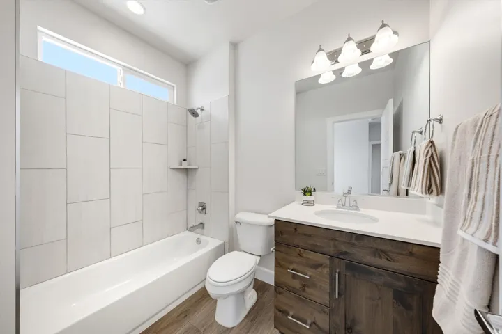 White subway-style tile shower surround with a built-in shelf, deep soaking tub, wood-finish flooring, dark wood vanity with a white countertop and undermount sink, and a multi-bulb vanity light fixture