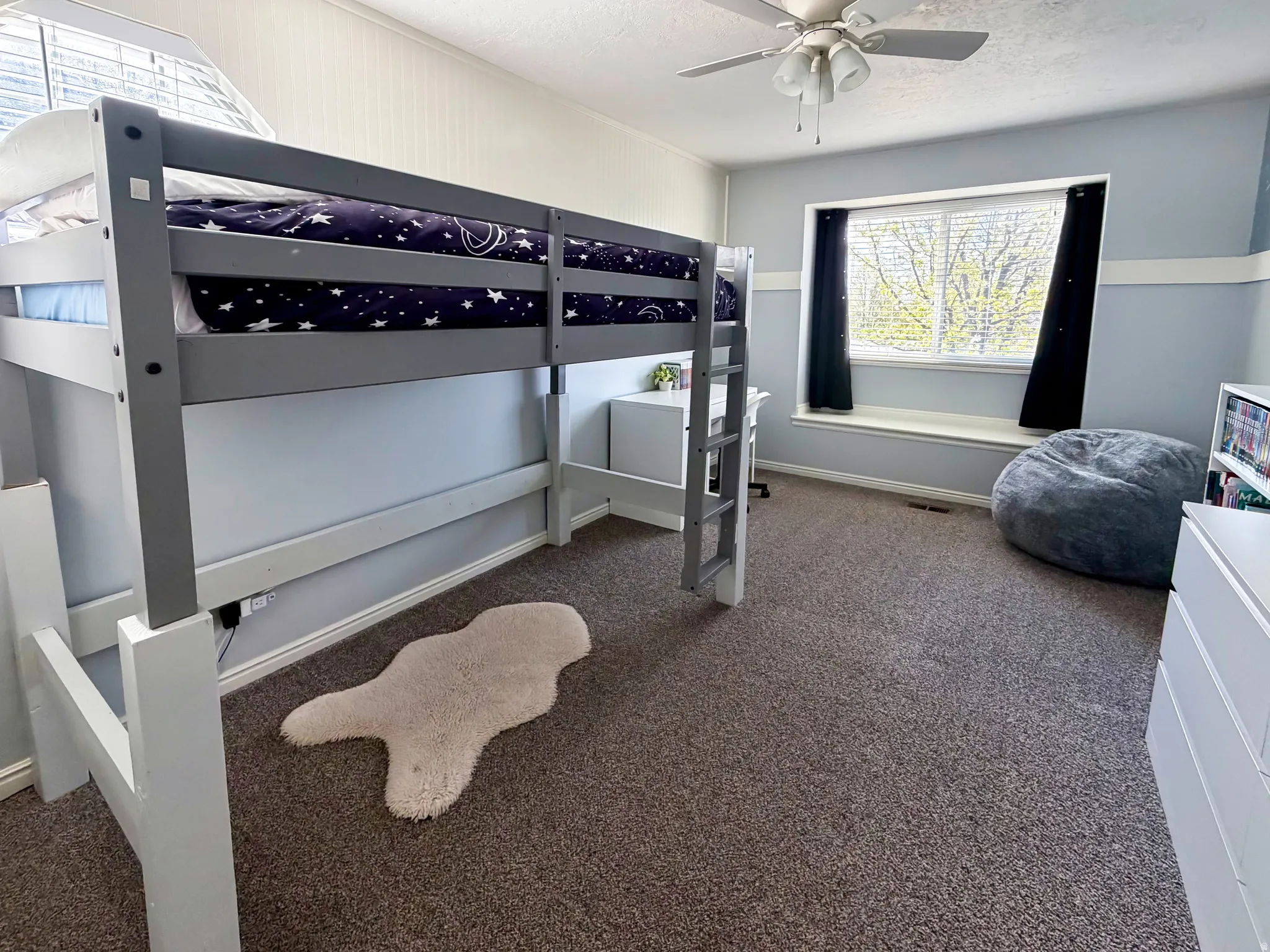 Built-in window seat, ceiling fan, carpeted flooring, and a dedicated desk space under the lofted bed frame