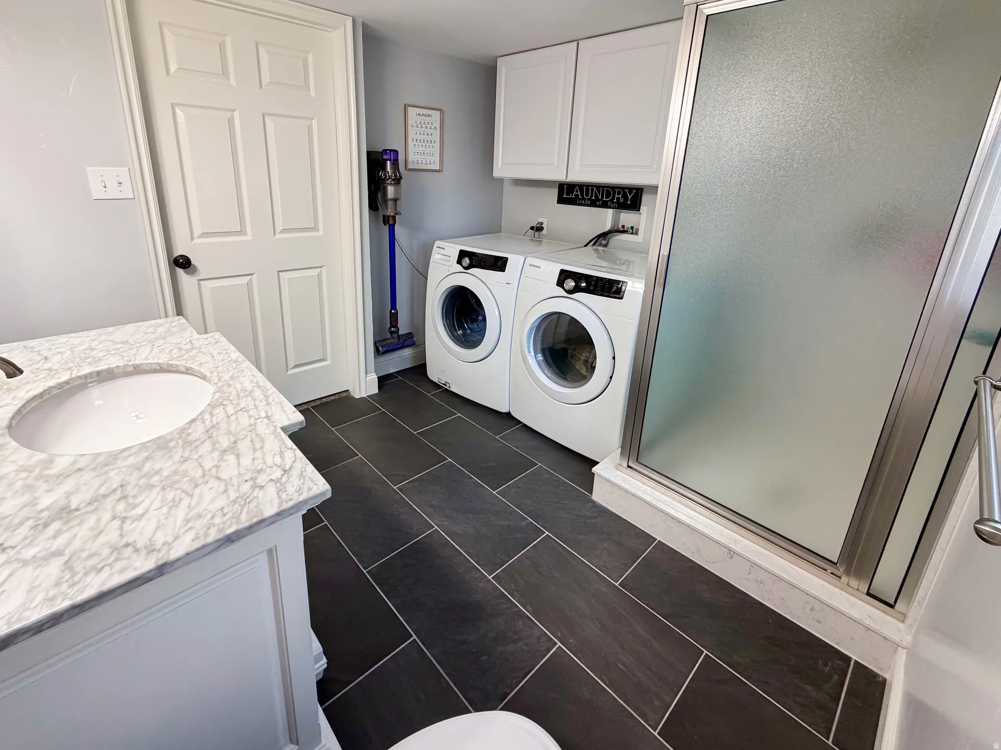 Vanity with integrated sink and stone-finish countertop, dark tile flooring, glass-enclosed shower, white cabinetry, and built-in laundry area