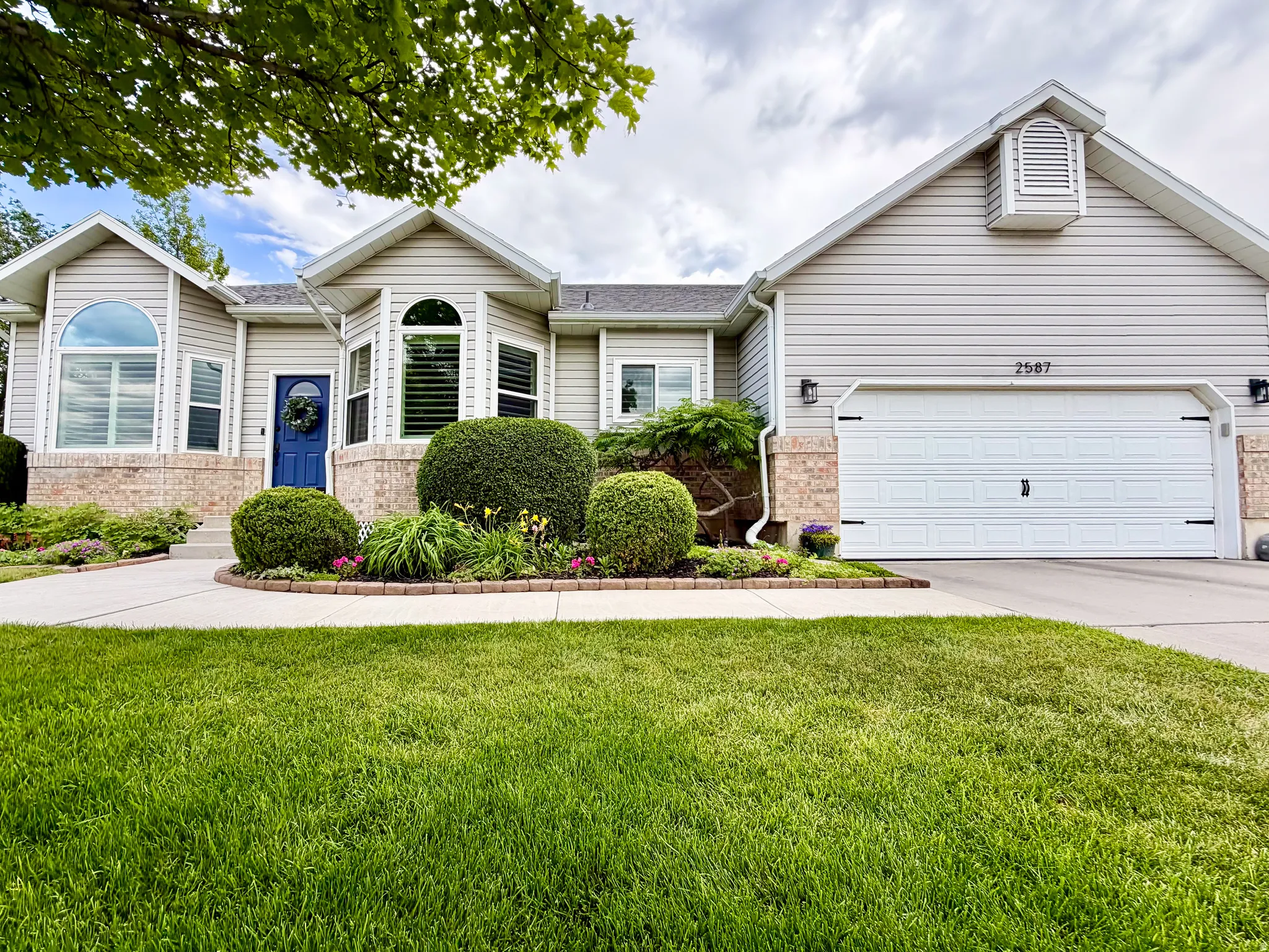 Traditional exterior with light-colored siding, brick accent detailing, a two-car garage, mature landscaping, and an arched entry window