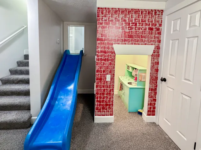 Carpeted stairs with white handrail, blue slide, brick-patterned accent wall, and built-in play kitchen with a sink