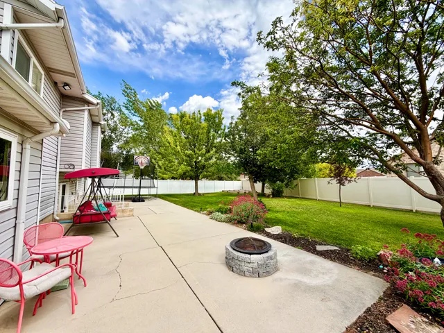 Expansive concrete patio with a built-in stone fire pit, bordered by a manicured lawn, established trees, and mature landscaping