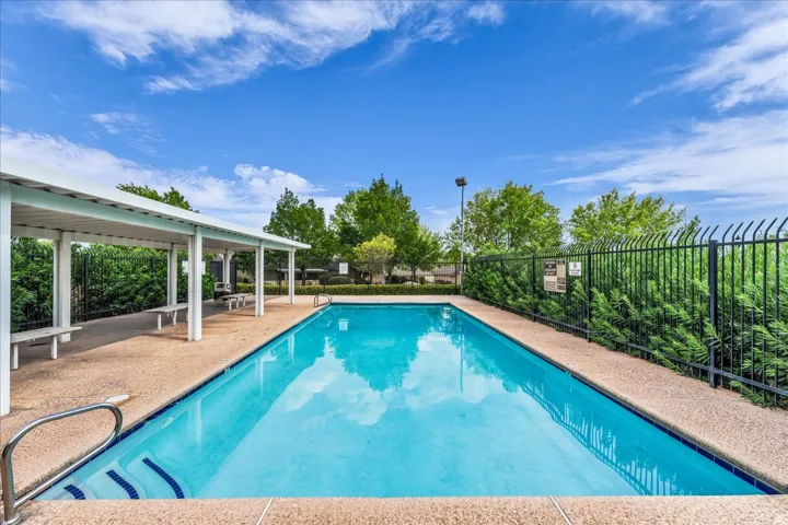 Rectangular swimming pool with integrated steps, surrounded by a textured concrete deck, a covered patio area with built-in benches, and mature perimeter landscaping