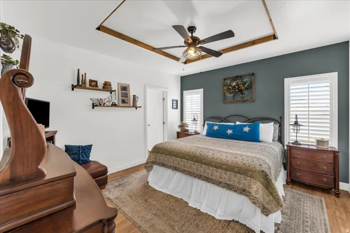 Wood-finish flooring, ceiling fan with light fixture, tray ceiling with wood beam accents, plantation shutters, and built-in wall shelving