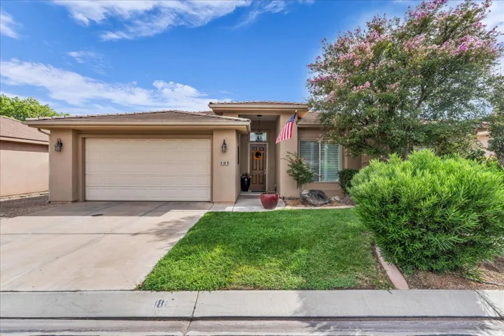 Welcoming single-story residence featuring a two-car garage with white paneled door, stucco exterior in warm neutral tones, and a manicured front lawn with mature landscaping including flowering shrubs and established greenery