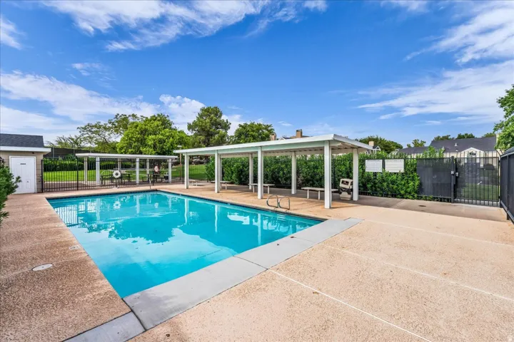Resort-style saltwater pool with surrounding concrete decking and mature landscaping, enclosed by a modern aluminum and glass pavilion structure offering shaded lounging and entertaining space