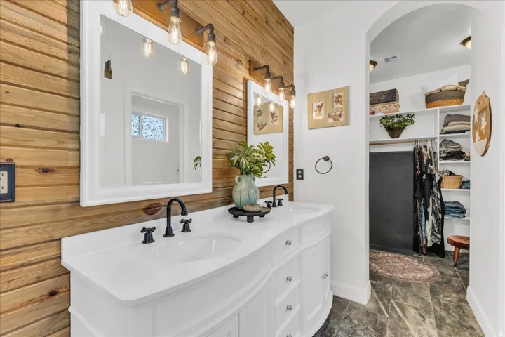Dual white vanities with integrated sinks, matte black fixtures, wood-finish wall paneling, and an arched entryway to a closet with built-in shelving