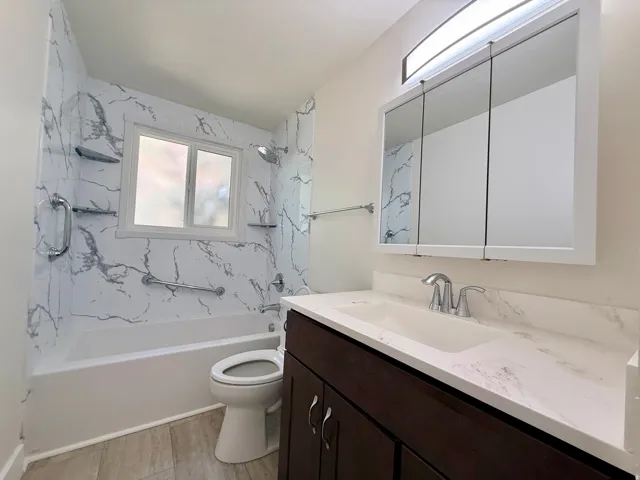 Bathroom featuring a shower/tub combination with white and gray patterned wall panels, a window above the tub, a vanity with a dark wood-finish cabinet and white countertop, and a mirrored medicine cabinet with integrated lighting
