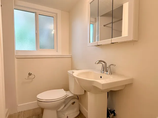 Pedestal sink with chrome faucet, wall-mounted medicine cabinet with mirrored doors, toilet, and frosted window