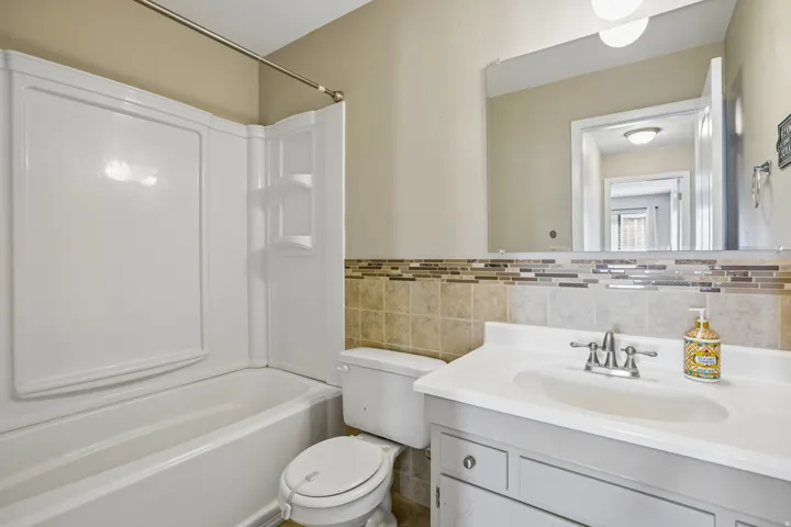 Bathroom featuring a single-sink vanity with white countertop, chrome faucet, tiled backsplash, wall-mounted mirror, and a full-size bathtub with shower surround
