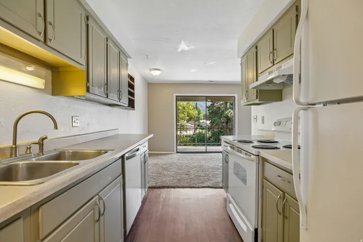 Galley kitchen featuring dual basin stainless steel sink, wood-finish flooring, ample cabinetry, and white appliances