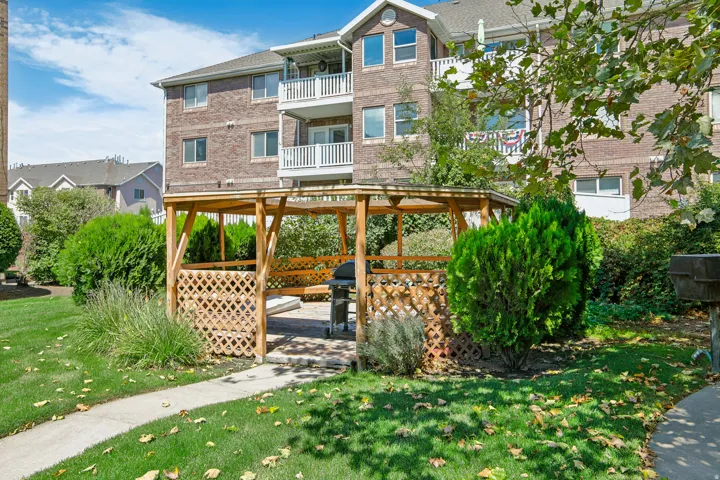 Brick exterior building with multiple balconies, wood pergola with lattice detailing, paved patio area, manicured lawn, and mature landscaping