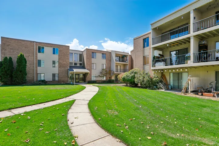 Expansive manicured lawn with curved concrete walkway, mature evergreen landscaping, and multi-unit residential buildings featuring brick and stone facades with balconies and ground-level patios