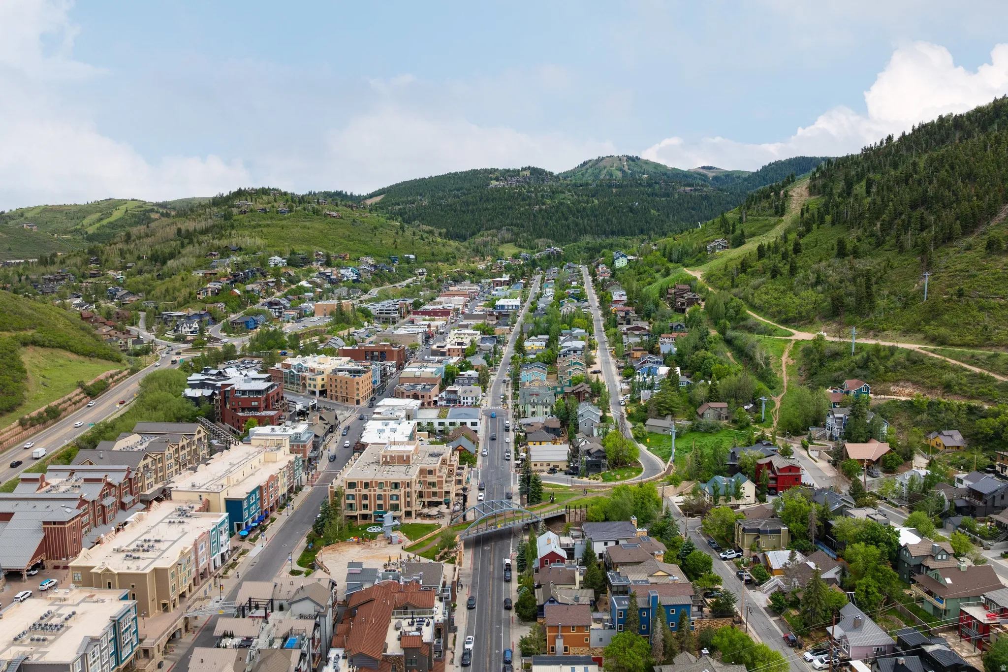 Townscape with diverse architectural styles, surrounded by lush green hills, with a prominent road network, and a distant mountain backdrop