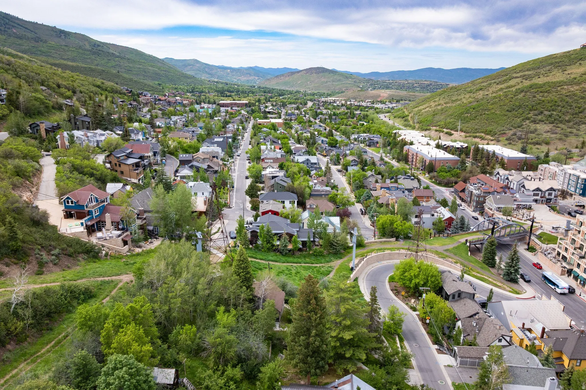 Community featuring diverse architectural styles, surrounded by green mountains and mature trees, with a winding road network and multiple residential structures