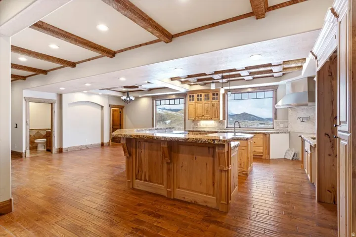 Open-concept kitchen and living area featuring wood-finish flooring, exposed ceiling beams, a central island with a granite countertop, wood cabinetry, and large windows revealing a mountain landscape