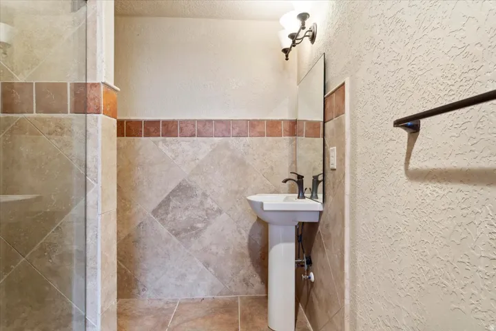 Pedestal sink with dark bronze faucet, wall-mounted mirror, decorative wall sconce, full-height tile shower enclosure, and coordinating tiled walls
