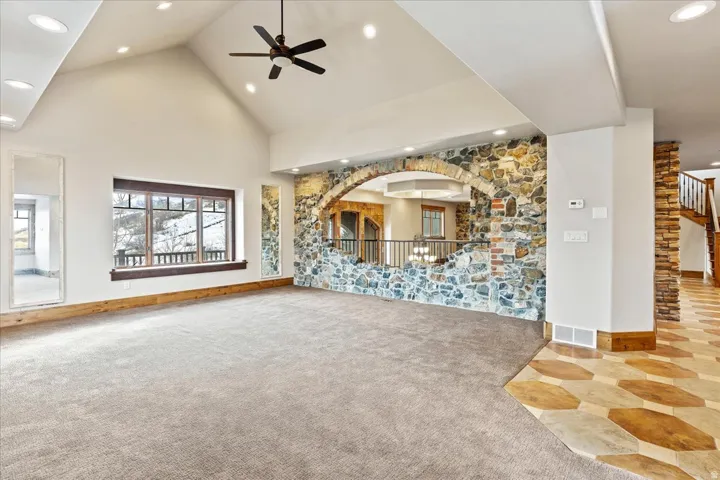 Vaulted ceiling with recessed lighting and ceiling fan, stone accent wall with arched opening, large picture window with wood trim, and wood-finish flooring