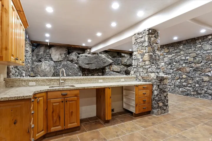 Stone-clad walls, recessed lighting, wood-finish cabinetry, a stone-finish countertop, and a stainless steel sink
