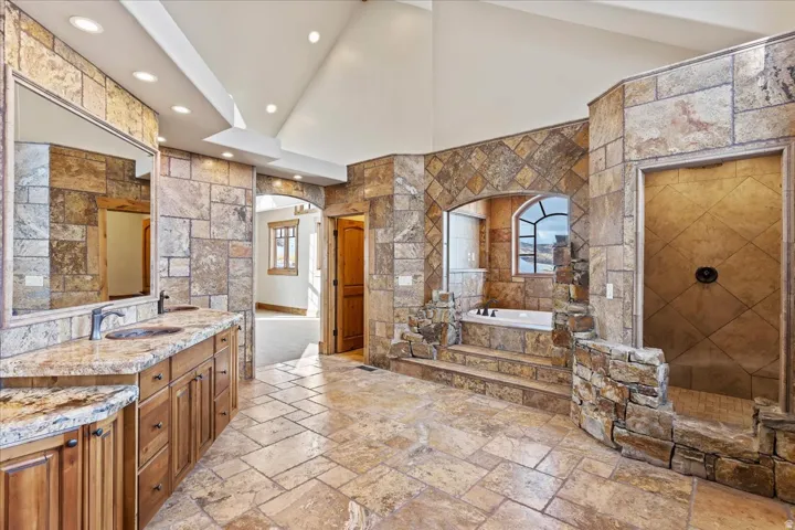 Expansive bathroom featuring a built-in soaking tub, stone-tiled walls, a walk-in shower, a dual vanity with granite countertop, and tiled flooring