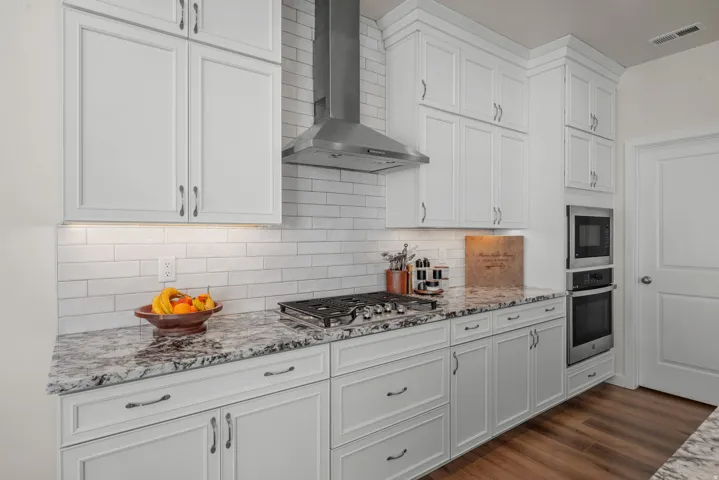White shaker-style cabinetry with dark hardware, stainless steel range hood, stone countertops, subway tile backsplash, and built-in stainless steel wall oven and microwave