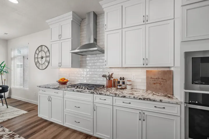White shaker-style cabinetry with dark hardware, stone countertops with natural veining, stainless steel range hood, white subway tile backsplash, and wood-finish flooring