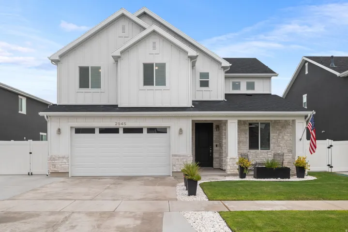Modern two-story residence featuring a clean-lined white exterior with mixed stone and fiber cement accents, a three-car garage with white paneled doors, and a professionally landscaped front yard with manicured lawn, decorative planters, and stone hardsc
