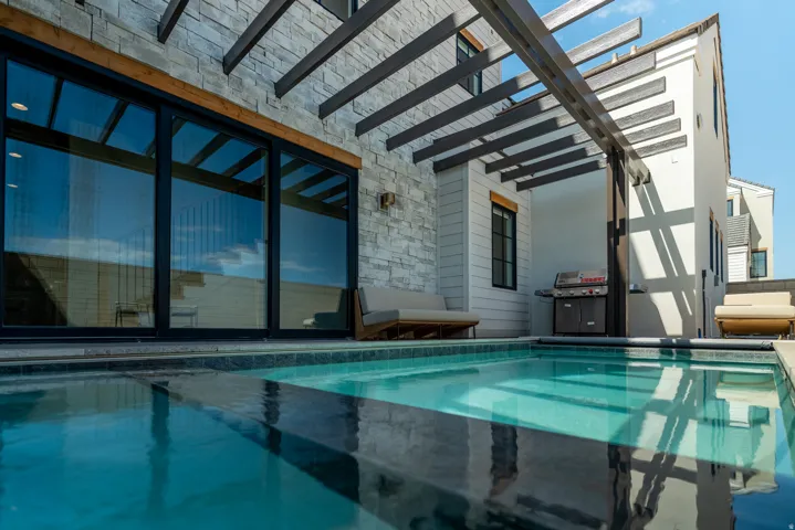 Outdoor swimming pool with integrated steps, stone-clad exterior wall, expansive glass doors, and a wood pergola