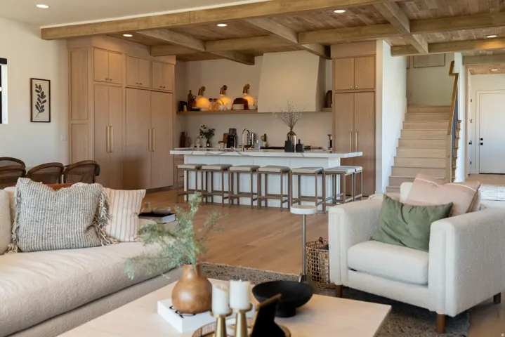 Open-concept living area featuring wood-finish flooring, a kitchen island with seating, integrated cabinetry, exposed wood ceiling beams, and a wood staircase with a dark metal handrail