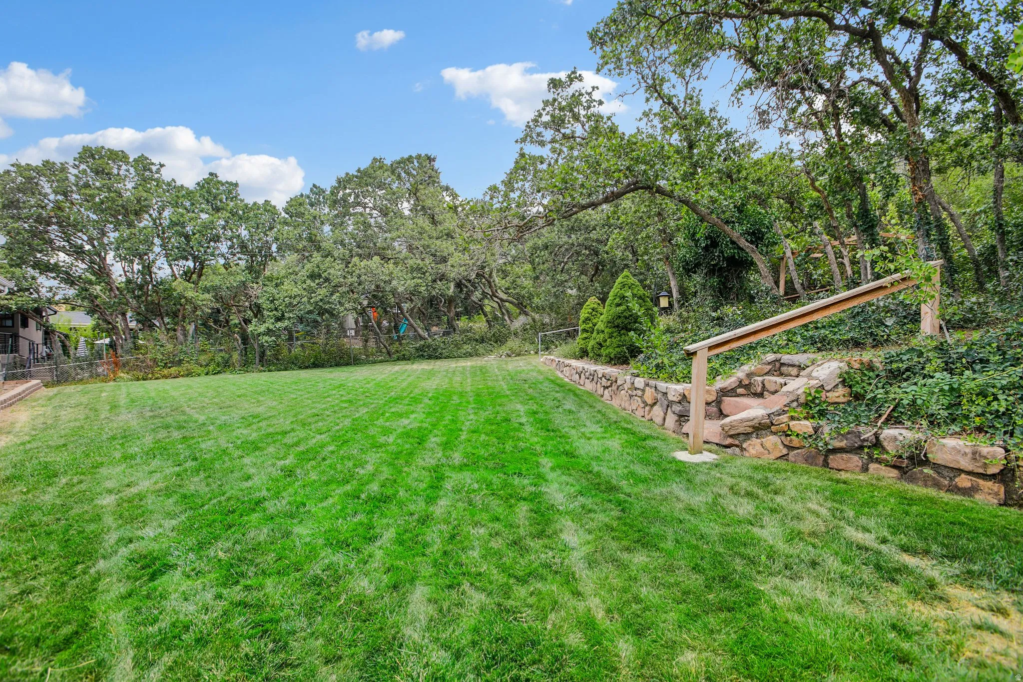 Expansive lawn with mature trees and a stone retaining wall featuring integrated stone steps and a wood handrail