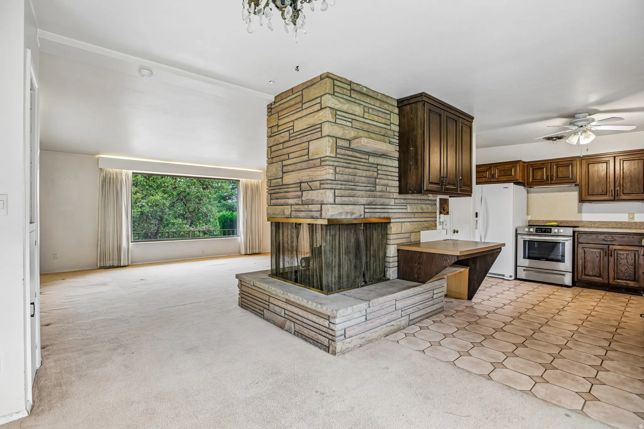 Stone-clad fireplace with hearth, kitchen with wood cabinetry, stainless steel range, and hexagonal tile flooring, and broad window overlooking mature trees