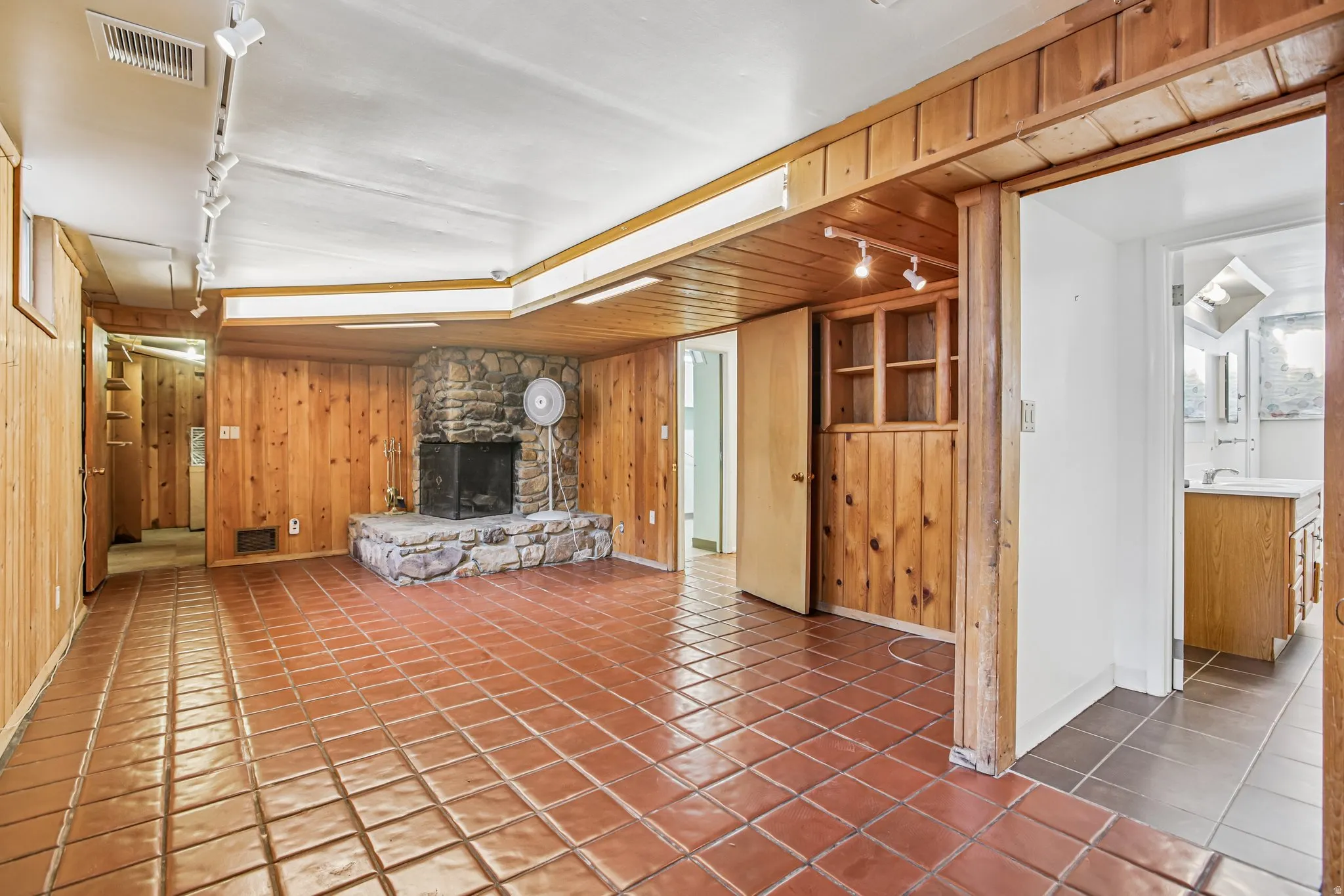Stone fireplace with hearth, wood-paneled walls, tile flooring, built-in shelving, and track lighting
