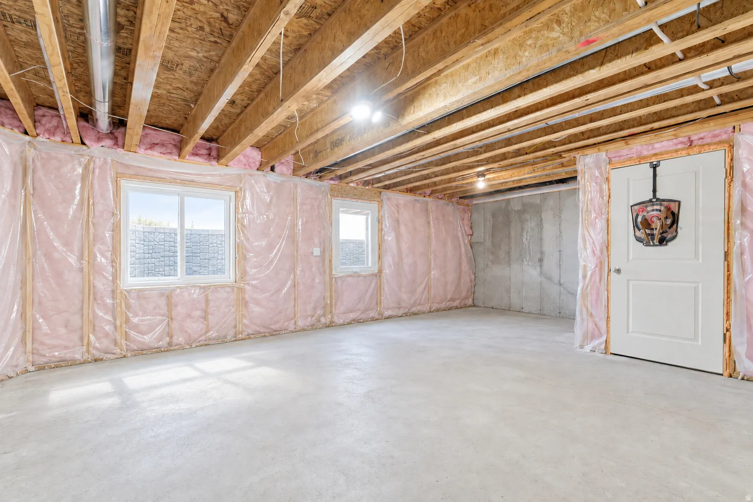 Unfinished basement with concrete flooring, exposed wood ceiling joists, two windows, insulated wall framing, and a white interior door