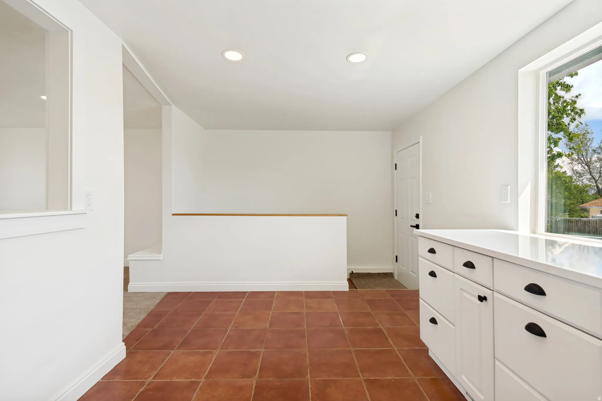 Terracotta tile flooring, white cabinetry with matte black hardware, white solid surface countertop, large window, and recessed lighting