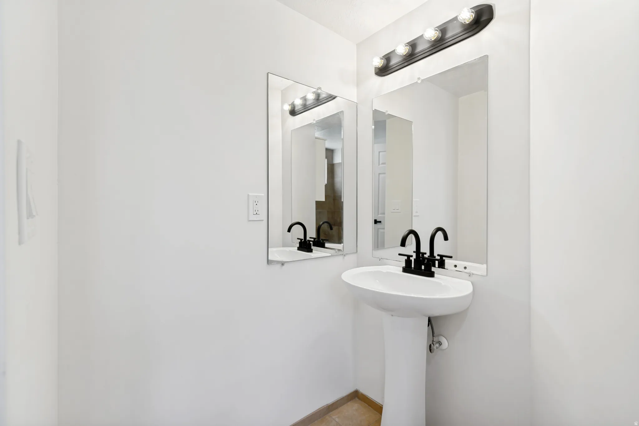 Pedestal sink with matte black faucet, wall-mounted mirror, and overhead vanity lighting