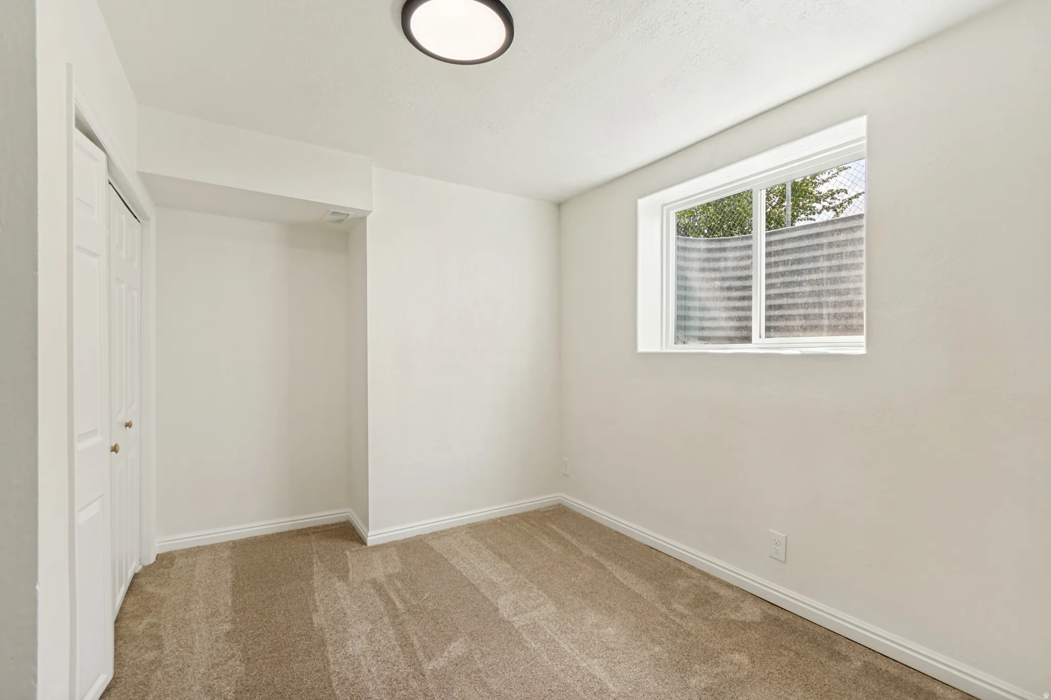 Neutral wall paint, carpeted flooring, window with exterior well, bi-fold closet doors, and a ceiling-mounted light fixture
