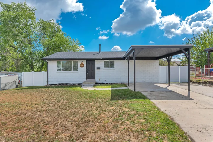 White painted brick exterior with a dark roof, covered carport, concrete driveway, and a white fence