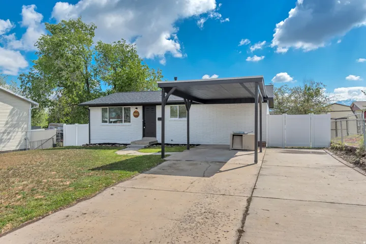 White painted brick exterior with a dark roof and a carport, concrete driveway, and a white fence