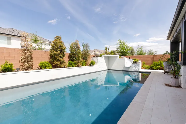 Rectangular swimming pool with integrated water features, expansive patio with tile pavers, landscaped beds, and a covered outdoor living area