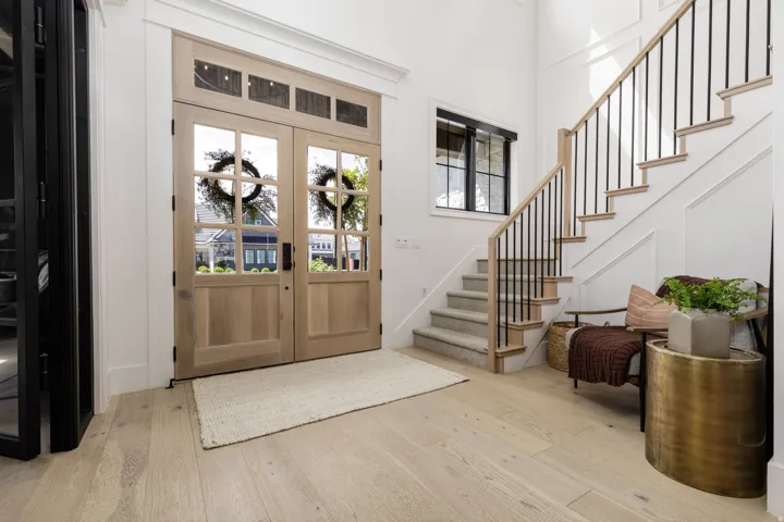Double entry doors with divided lites and transom windows, light wood-finish flooring, staircase with wood handrails and black metal balusters, white wall paneling, and a black-framed window